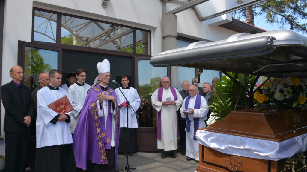 Catholic funeral outside a building with a bishop in purple vestments speaking at a microphone beside a decorated casket.