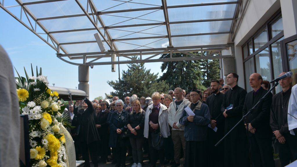 Crowd gathered under a glass-covered walkway for a funeral, with a large flower wreath in the foreground and clergy in black robes among attendees.