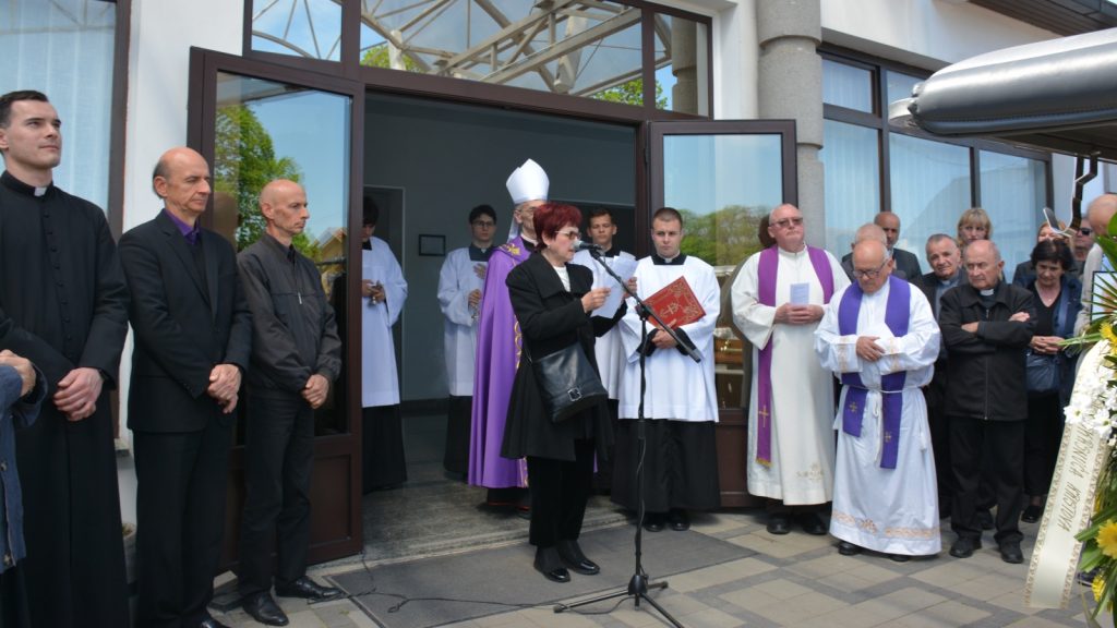 Group of clergy and attendees standing outside a church entrance as a woman speaks at a microphone.