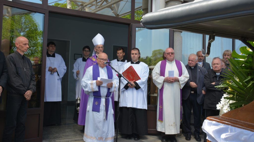 Catholic funeral outside a church entrance with priests in white and purple vestments leading prayers while a coffin is visible near a flower arrangement.