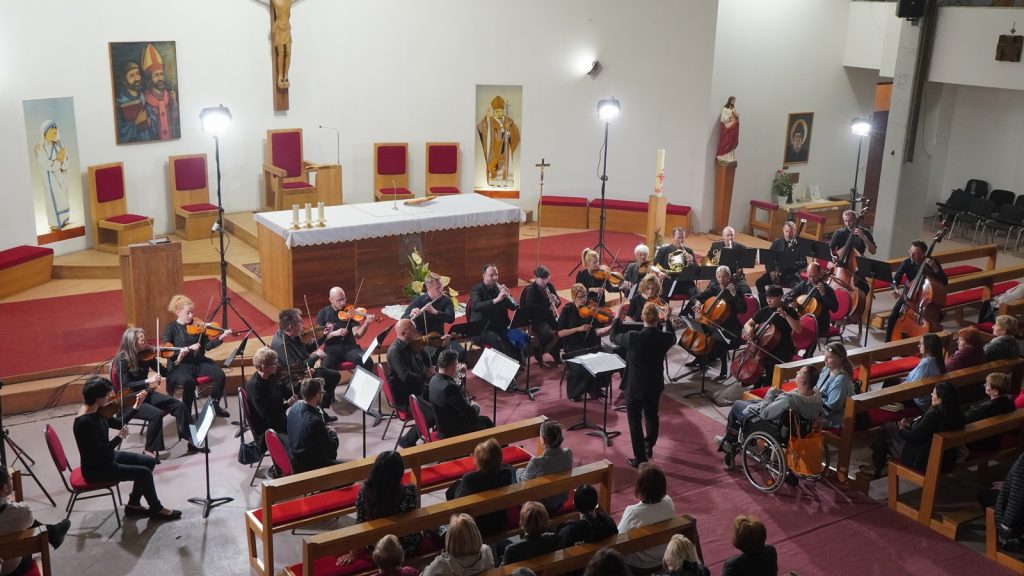 Orchestra performs in a church sanctuary with a conductor, seated string players, and an audience in pews; altar and religious statues in the background.