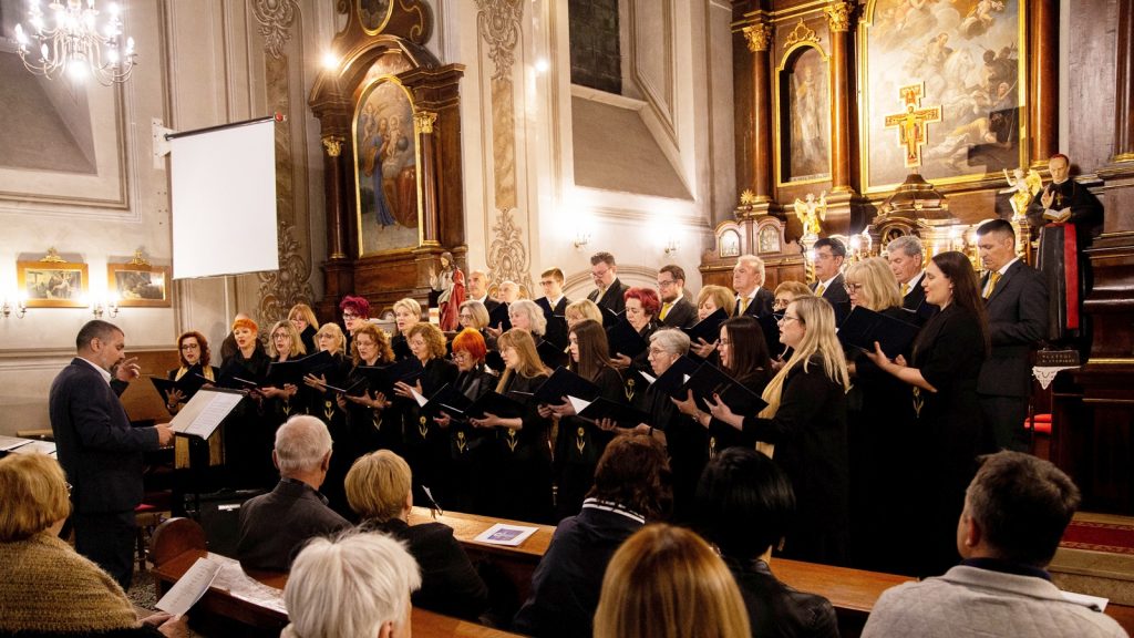 Choir wearing black robes performs in a ornate church with a conductor at left and an audience watching from pews.