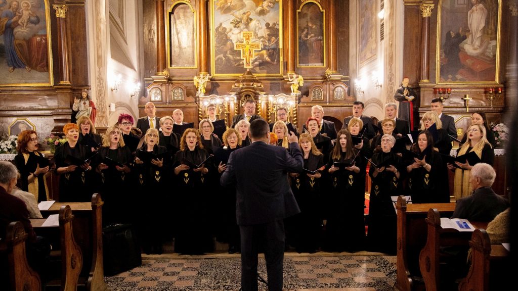 A choir in black robes performs in a ornate church, conductor facing them with raised hands amid pews and spectators in the foreground.