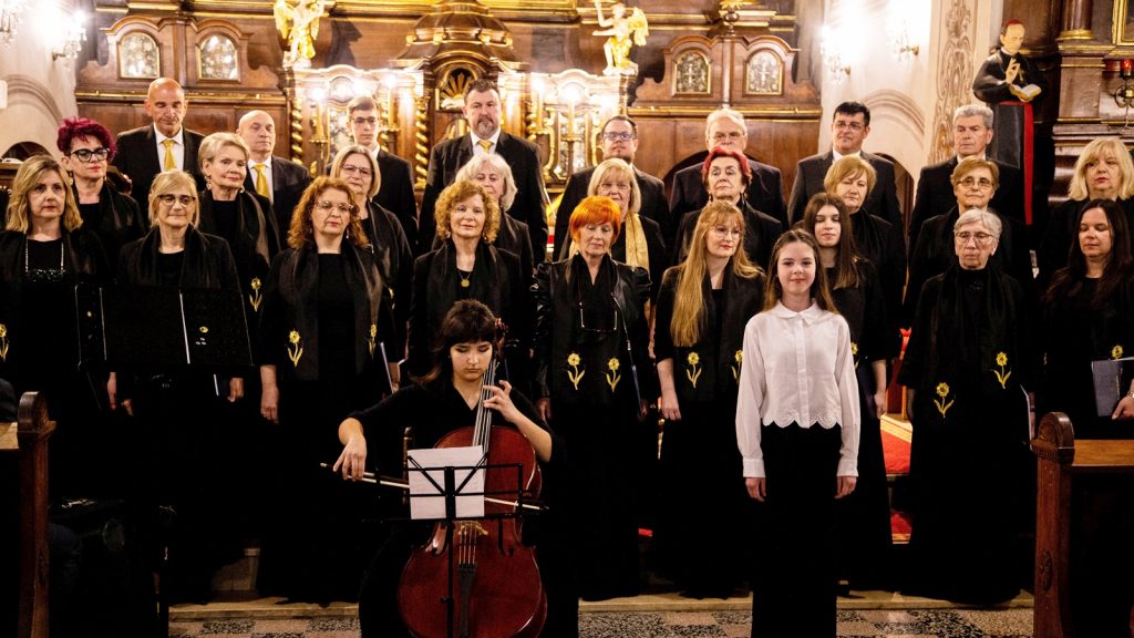Choir in black robes with gold flower insignias, standing in a decorated church sanctuary, with a young cellist foreground left and a girl in white front right.