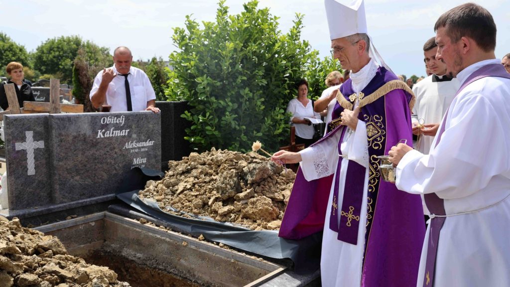Catholic priests in purple vestments blessing an open grave as mourners stand nearby at a cemetery.