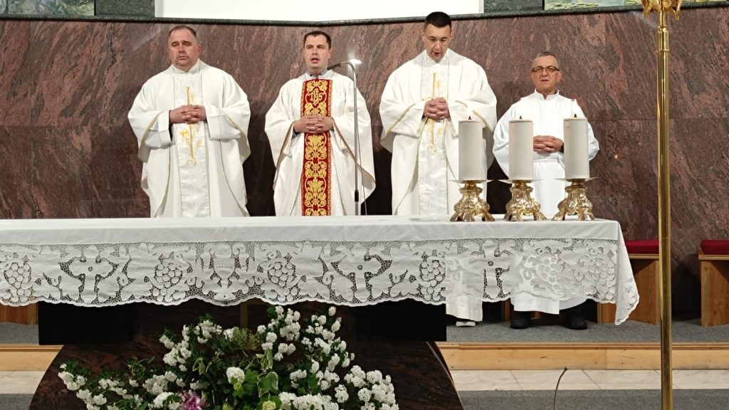 Four priests in white liturgical robes stand behind an altar with a white lace cloth during a church service, flanked by two tall candles and floral arrangements below.