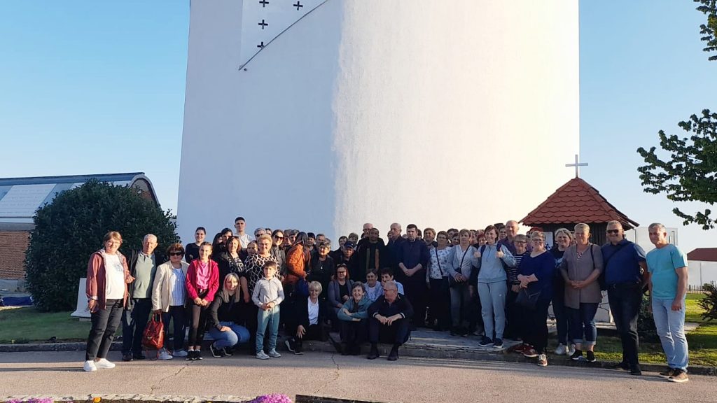 Group of people posing for a photo in front of a tall white tower with a small red-roofed structure and cross, on a sunny day.