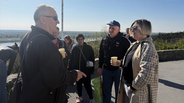 Group of adults standing outdoors on a sunny day, listening to a man in sunglasses who is speaking and gesturing with his hand inspecting the group.