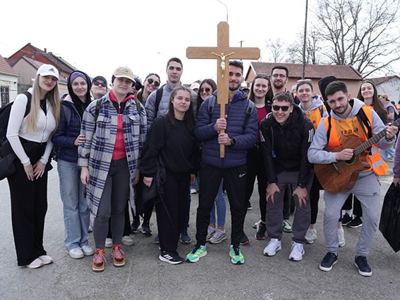 Group of young people posing on a street, a large wooden cross held by a central figure, guitarist on the right.