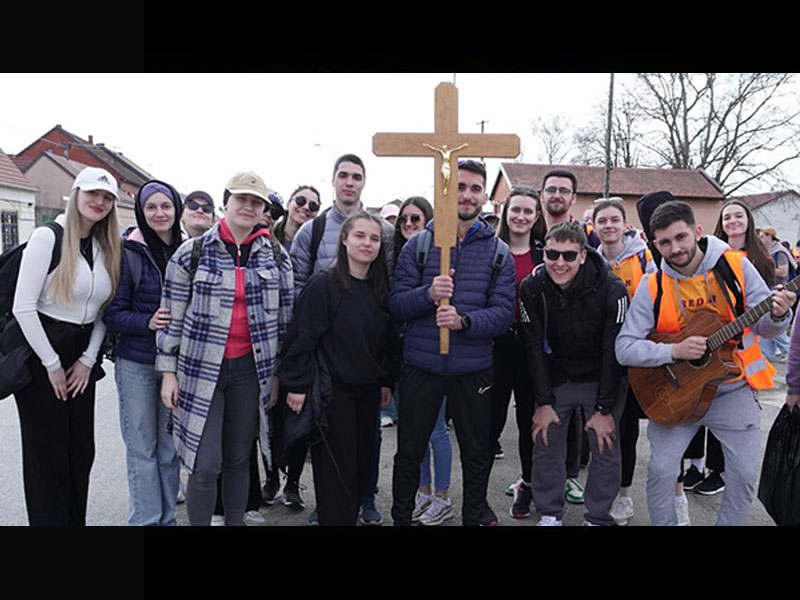Group of smiling young people outdoors, center holding a wooden cross, one person strumming a guitar on the right.