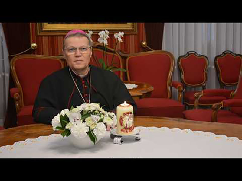 Catholic bishop in black vestments sits at a wooden table with a floral centerpiece and a lit candle, in a formal room with red upholstered chairs in the background.