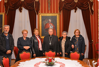 Six adults stand side by side in a formal dining room with red drapes and framed portraits; a round table with a floral centerpiece is in the foreground.