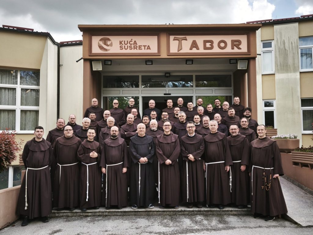 Large group of monks in brown robes standing on steps outside a monastery entrance.