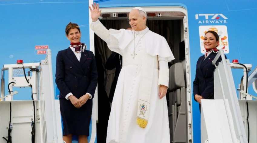Pope Francis waves from the doorway of a blue ITA Airways plane, flanked by two smiling flight attendants in navy uniforms.