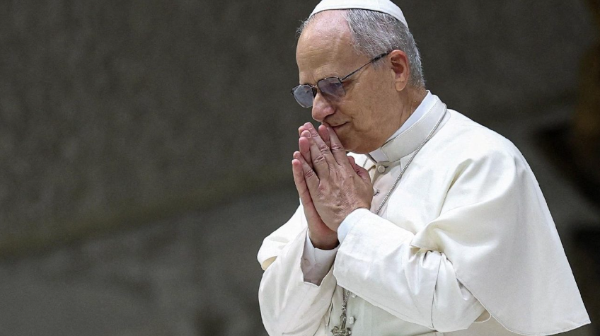Senior Catholic clergyman in white robes praying with hands clasped and head bowed.