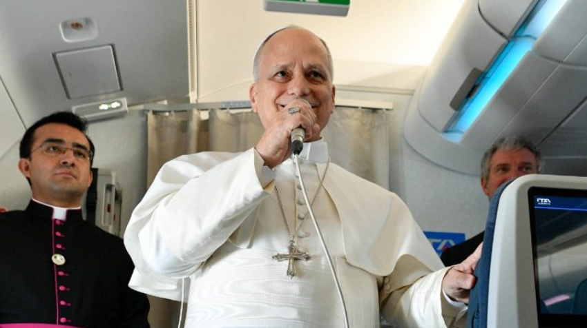 Catholic cleric in white robes speaks into a microphone on a plane, with another clergyman in dark attire nearby.