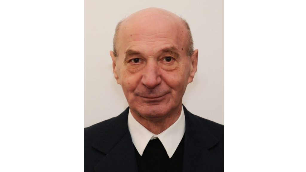 Portrait of an older man in a dark suit and white shirt, facing the camera against a light neutral background.