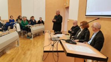 Panel discussion in a conference room with five speakers seated at a table and a presenter standing, addressing an audience.