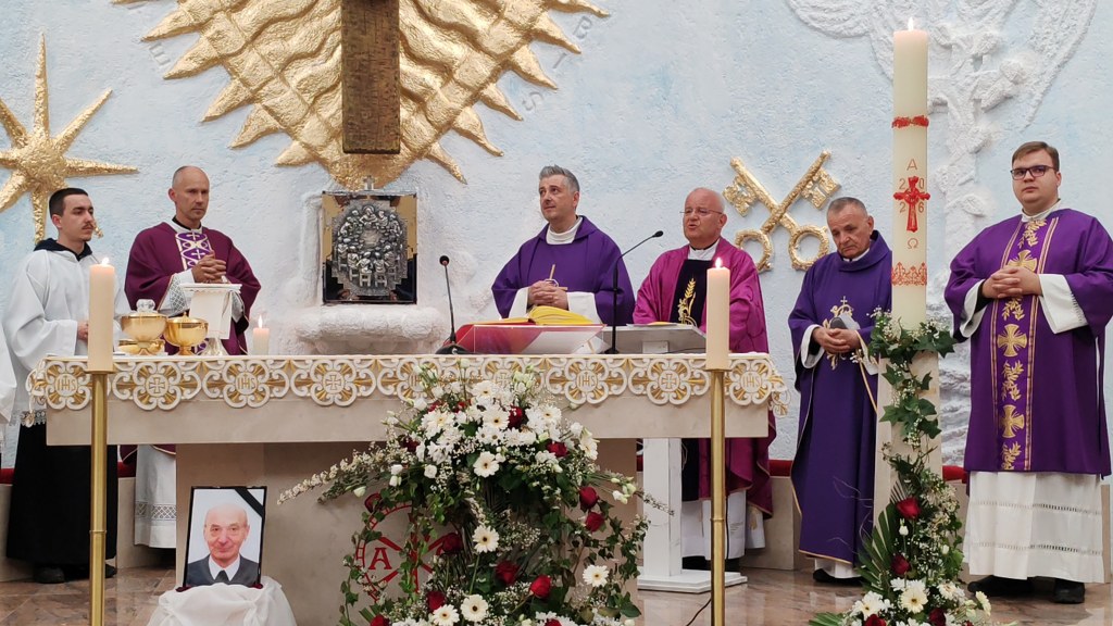 Catholic altar scene: six priests in purple vestments around a decorated altar with candles, flowers, and a framed portrait on the floor."