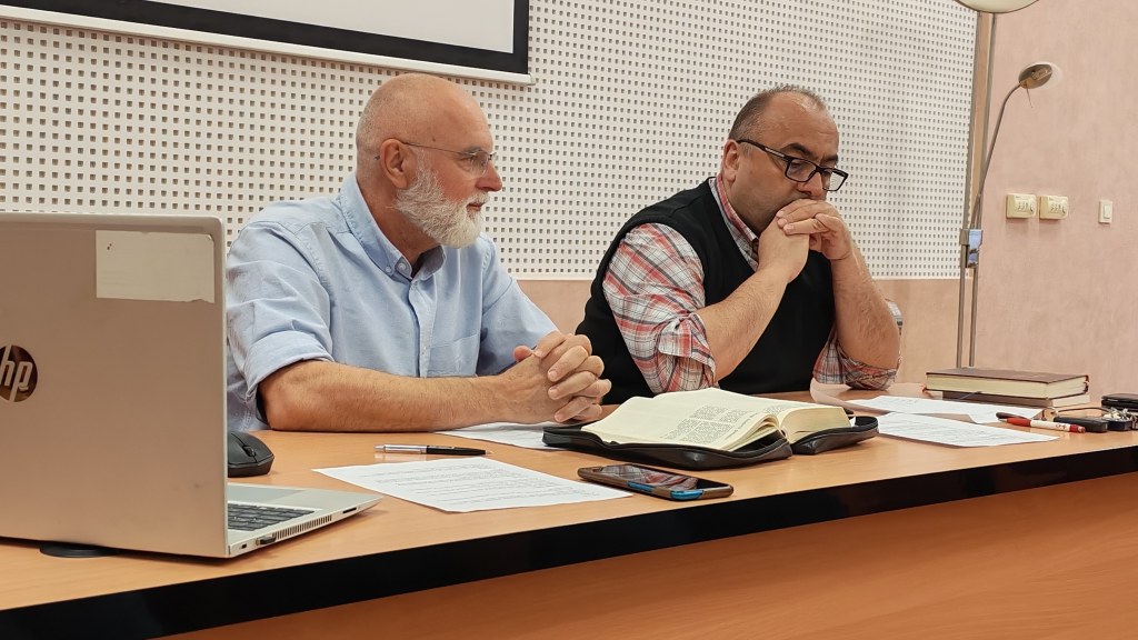 Two men sit at a conference table with papers and a large open book, one in a light blue shirt and the other in a plaid shirt and vest, engaged in discussion.
