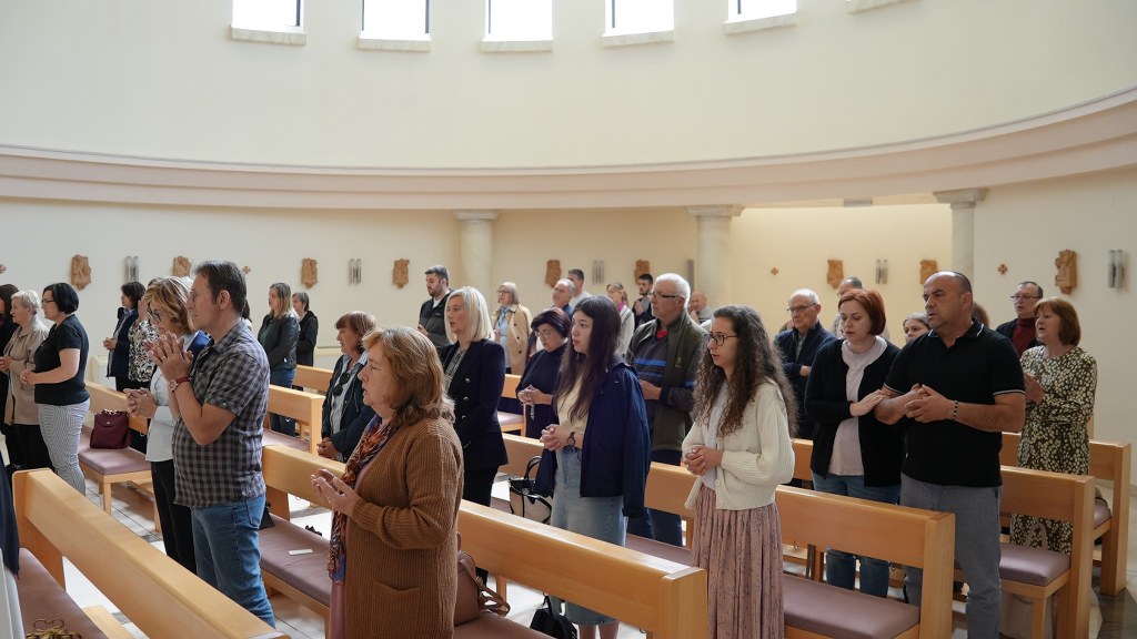 Group of people standing in a sunlit church, hands clasped in prayer beside wooden pews.