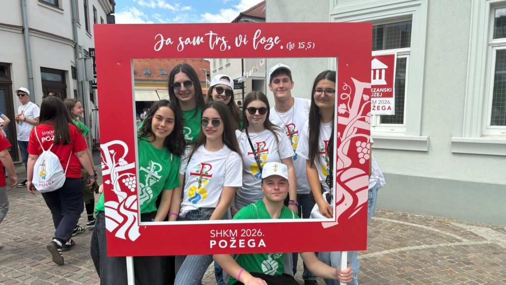 Group of smiling teens posing behind a large red photo-frame prop that says SHKM 2026 Požega at a street festival.