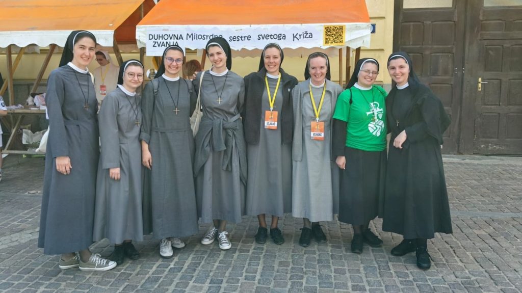 Eight nuns in gray habits stand together smiling outdoors, one woman in a green event shirt among them, on a cobblestone square in front of an orange tent stall.