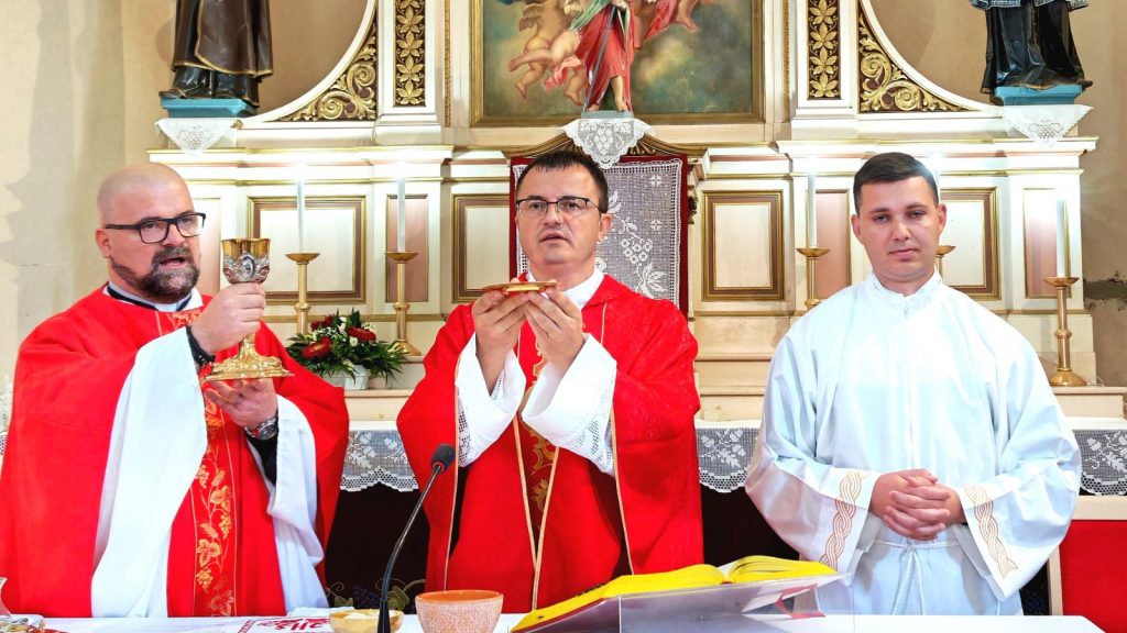 Three clergy at an ornate altar during Mass: left priest holds a gold chalice, center priest elevates the Eucharist, right altar server in white vestments.
