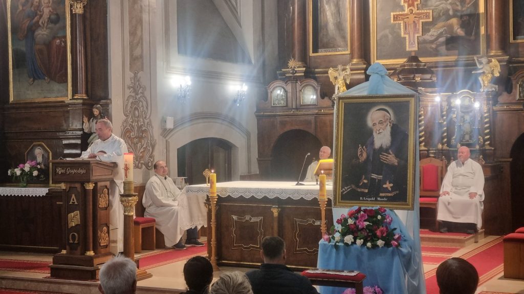 Priest at lectern delivering sermon inside ornate church, with altar and large framed portrait of a bearded saint nearby.