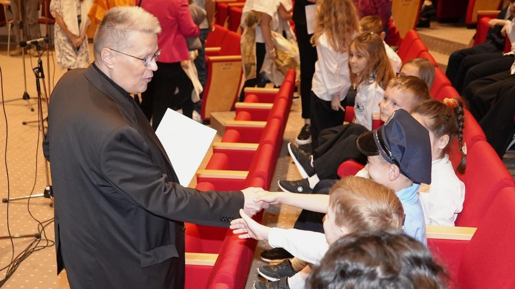 A man in a dark suit shakes hands with excited children seated in red chairs at an event or ceremony in a auditorium.