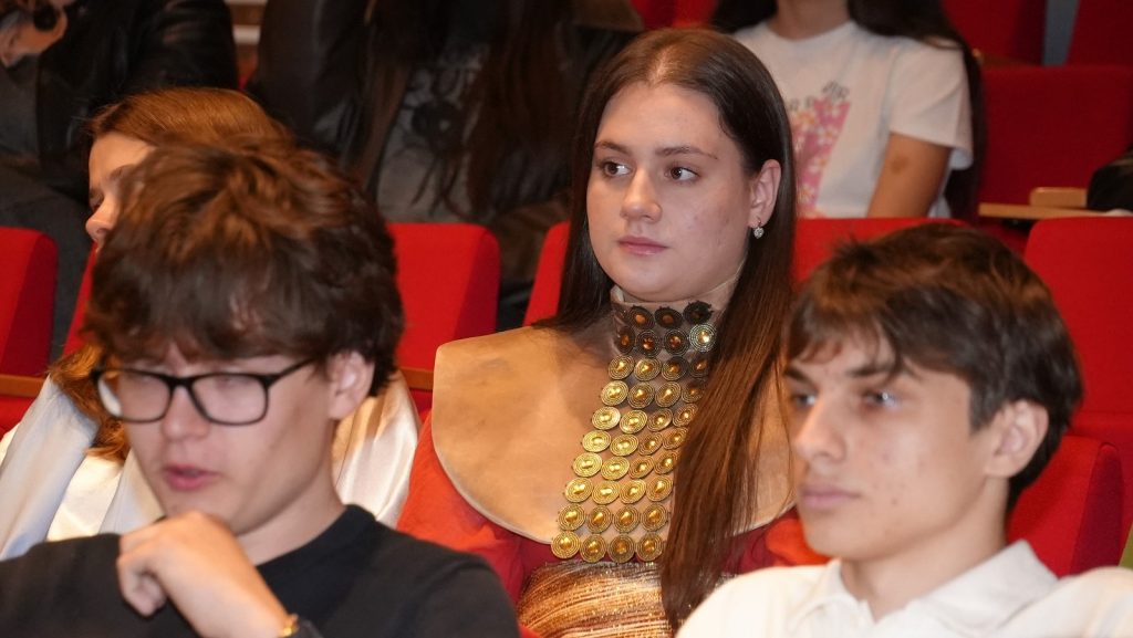 Young woman with a distinctive gold circular neckpiece sits in a red-carpeted theater seating area, looking alert to the side.