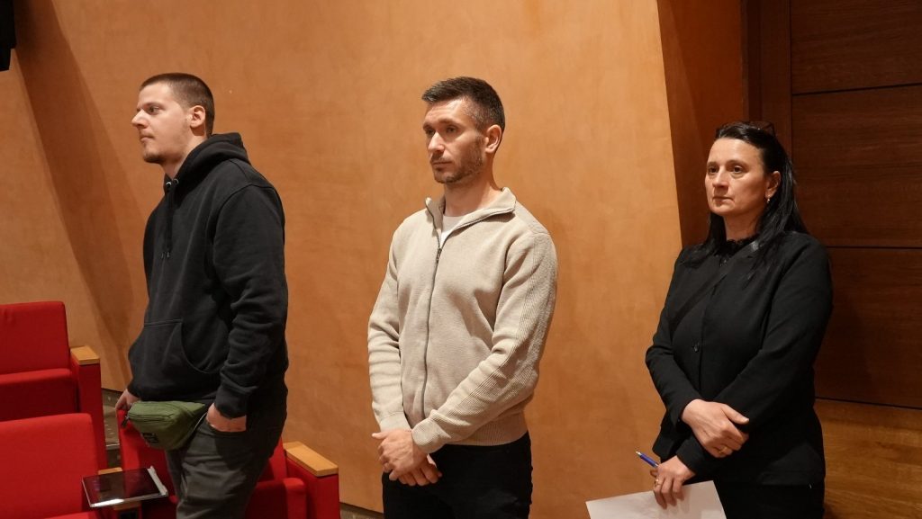 Three adults stand side by side in a theater or conference room, with red seats and wooden walls behind them.
