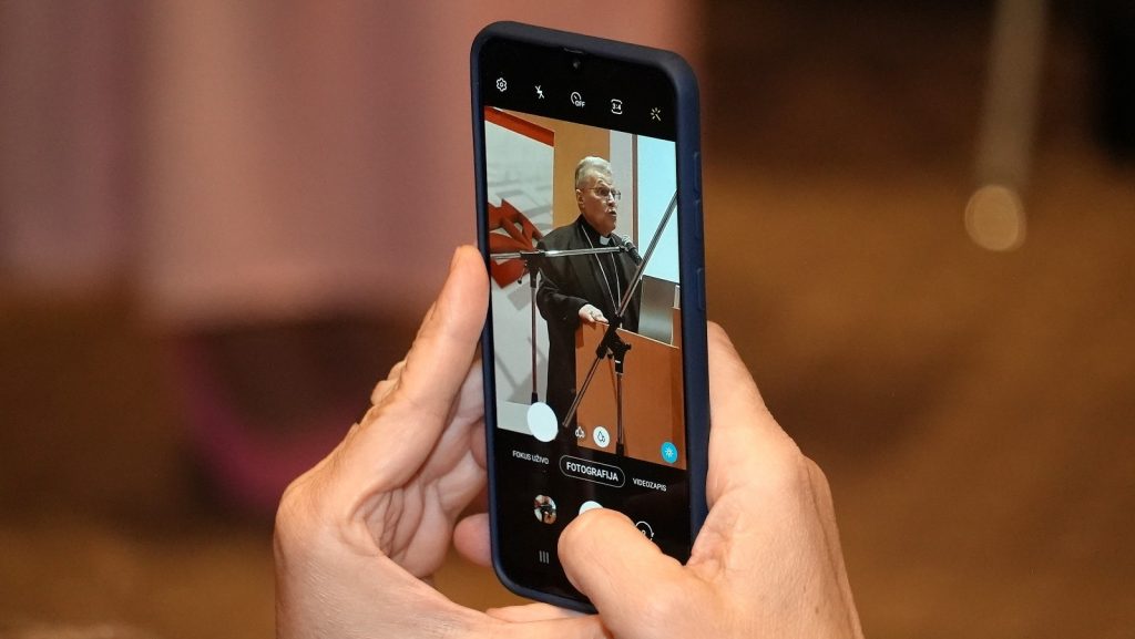 Person holds a smartphone upright, capturing an older man speaking at a podium with a microphone in a formal event setting