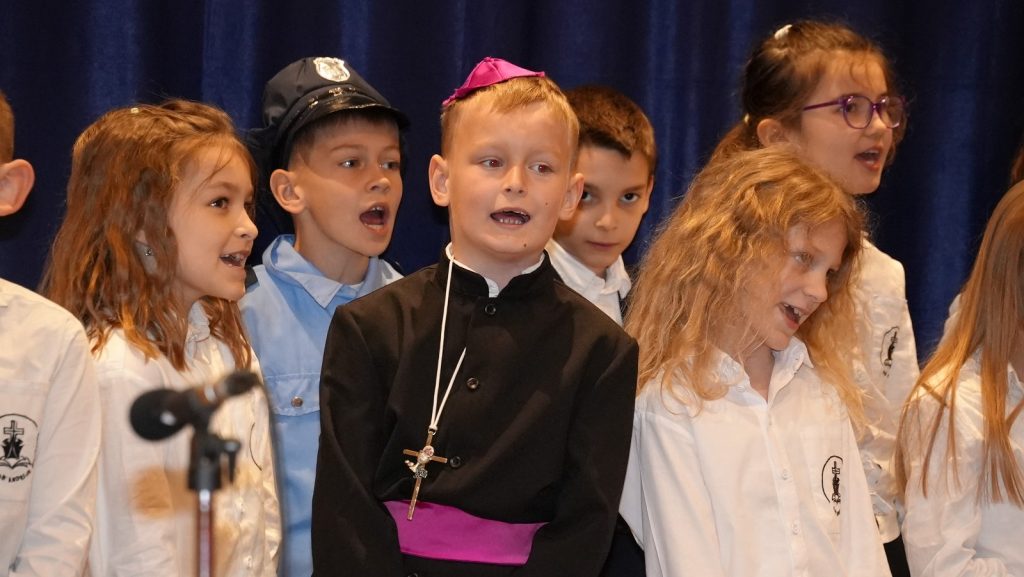 Group of schoolchildren singing on stage, center boy in black cassock with pink sash and cross necklace, others in white shirts against a blue curtain.
