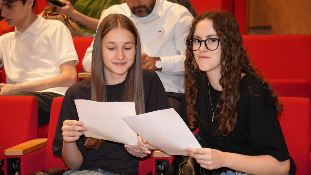 Two young women sit in red auditorium seats, looking at printed documents together.