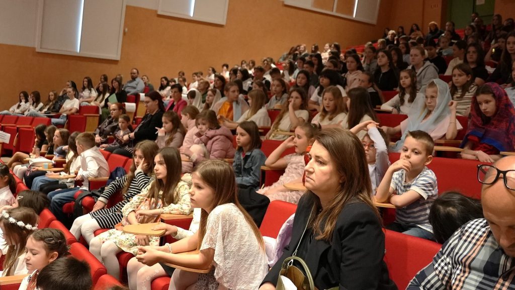 Crowded auditorium filled with children and adults watching a performance from red seats.