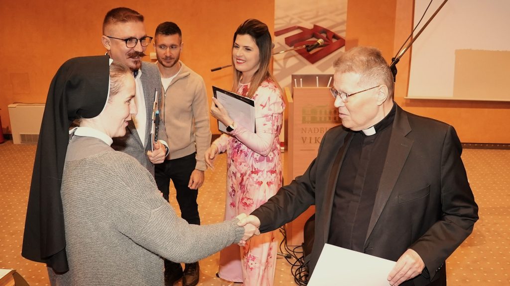 Priest in a black suit shakes hands with a nun in a gray sweater during an event while others watch.