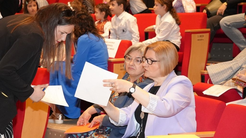 Seated woman in a lavender blazer shows a document to a standing woman with long brown hair in a red-seated auditorium.