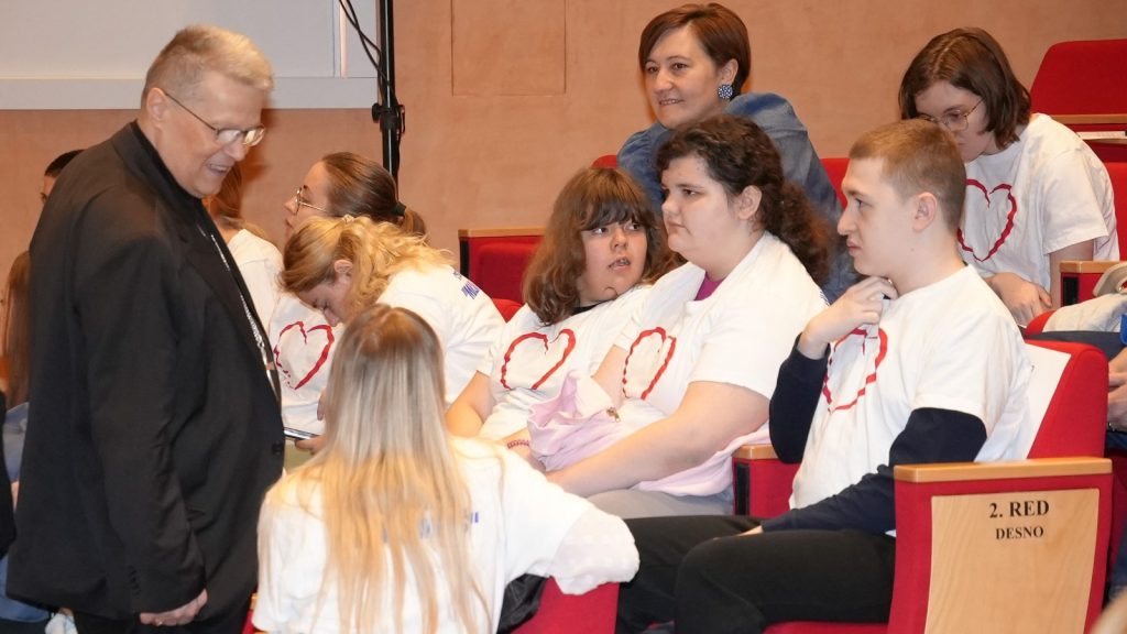 Man in a black jacket chats with a group of youths wearing white T-shirts with red heart logos in an auditorium seating area.
