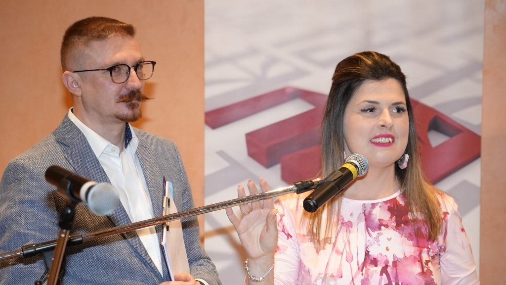 Woman in a pink floral top speaks into a microphone while a man in a blazer listens beside her, on a stage with a backdrop.