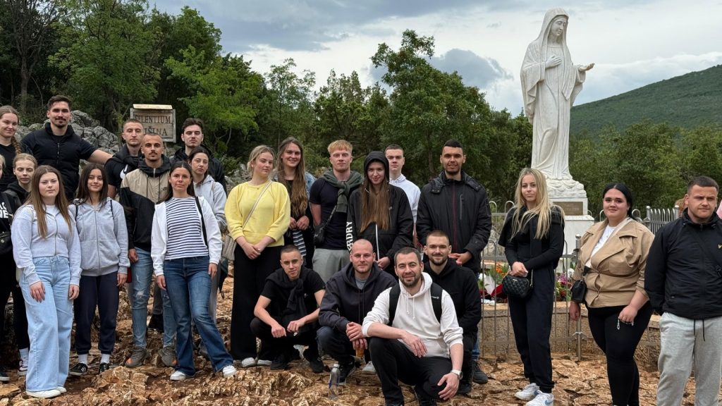 Group of young adults posing for a photo on rocky ground with a large white statue of the Virgin Mary in the background, outdoors near greenery.