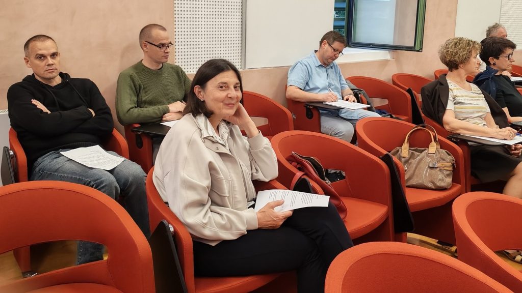 Group of adults seated in orange auditorium chairs, some taking notes and holding papers; woman in foreground smiles at camera.