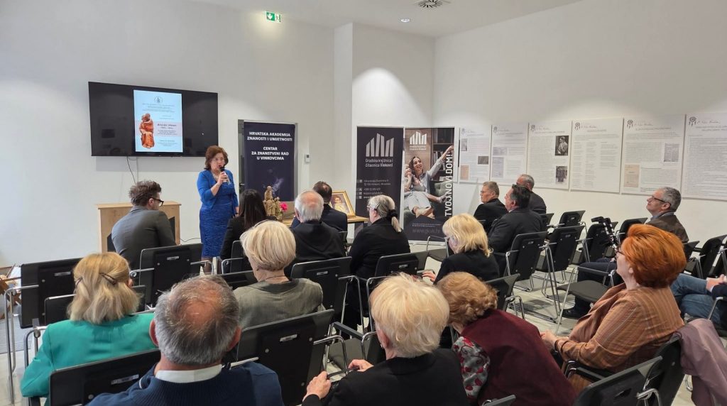 Woman in blue dress speaking to an audience in a bright conference room with banners and a TV screen.