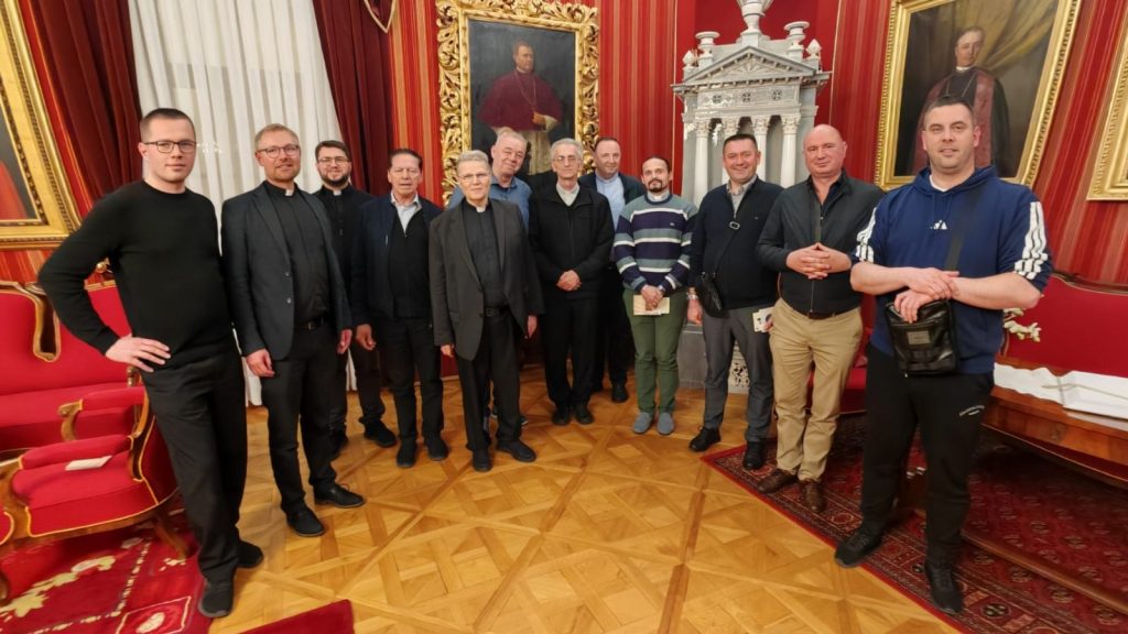 Group of men, including several clergy in clerical collars, posing in an opulent room with red walls, gold-framed portraits, and ornate decor.