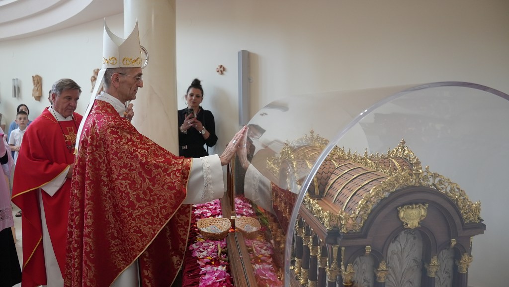 Catholic bishop in red vestments touches a gilded reliquary inside a glass case as attendants observe nearby.