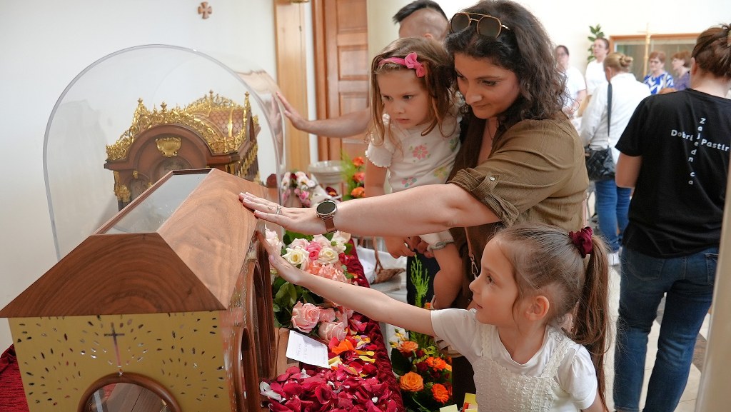 Mother and two young girls touch a glass-covered ornate reliquary in a flower-adorned display area.