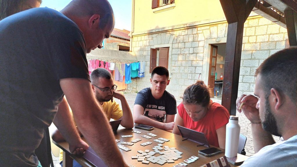 Group of people around a wooden table playing a tile-based board game outdoors, with laundry hanging on a line in the background.
