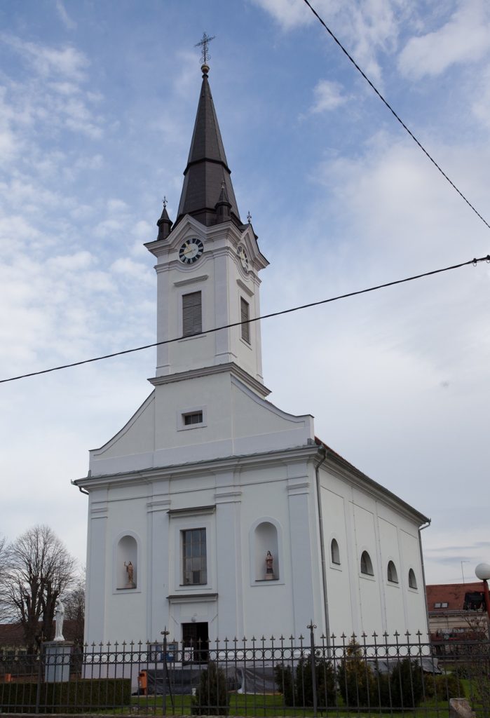 White church with a tall clock-tower steeple against a blue sky, fenced yard in front and statues in niches along the walls.