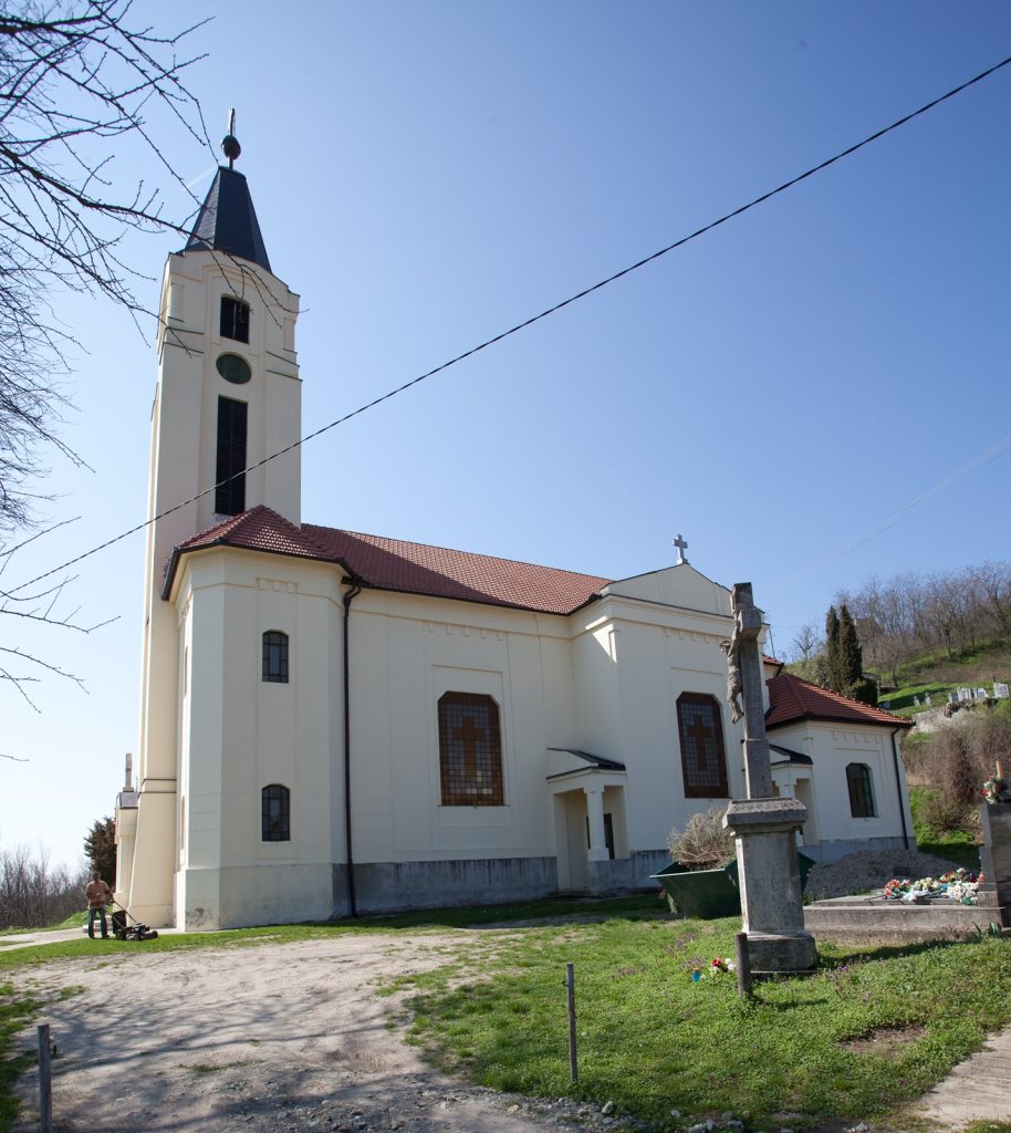 White church with a tall steeple and red-tiled roof against a clear blue sky, with a small grave marker in the foreground.