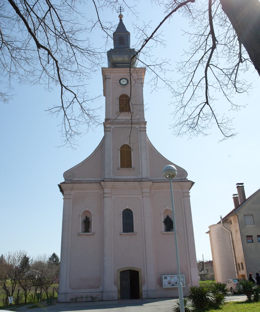 Front view of a pale church with a tall clock tower, framed by bare tree branches against a clear blue sky.